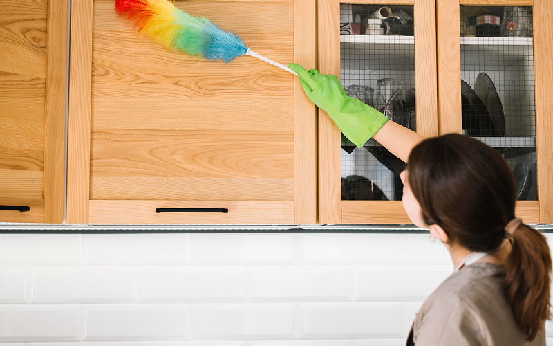 Domestic helper dusting wooden kitchen cabinets using colorful duster.