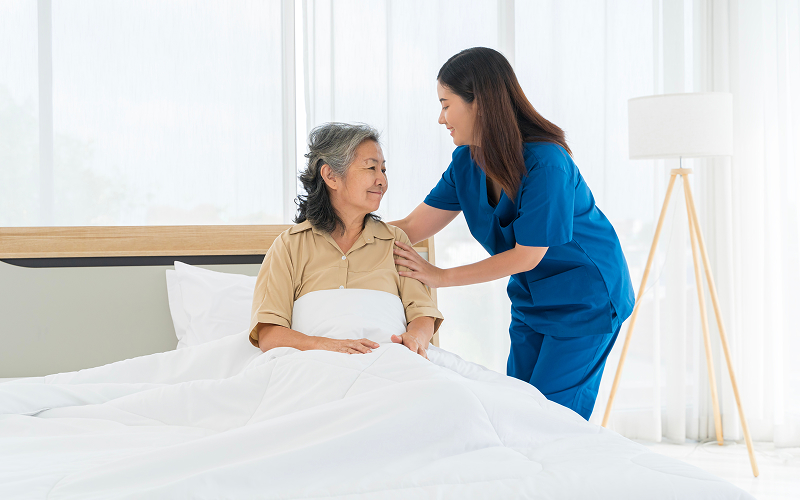 Experienced caregiver assisting elderly woman sitting on bed.